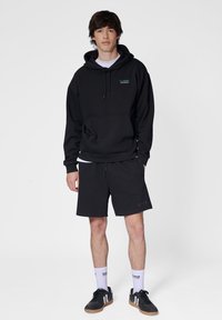 Zwarte pullover hoodie met een voorzak, gecombineerd met zwarte shorts. Model draagt een witte t-shirt eronder, zwarte sneakers en witte sokken.
