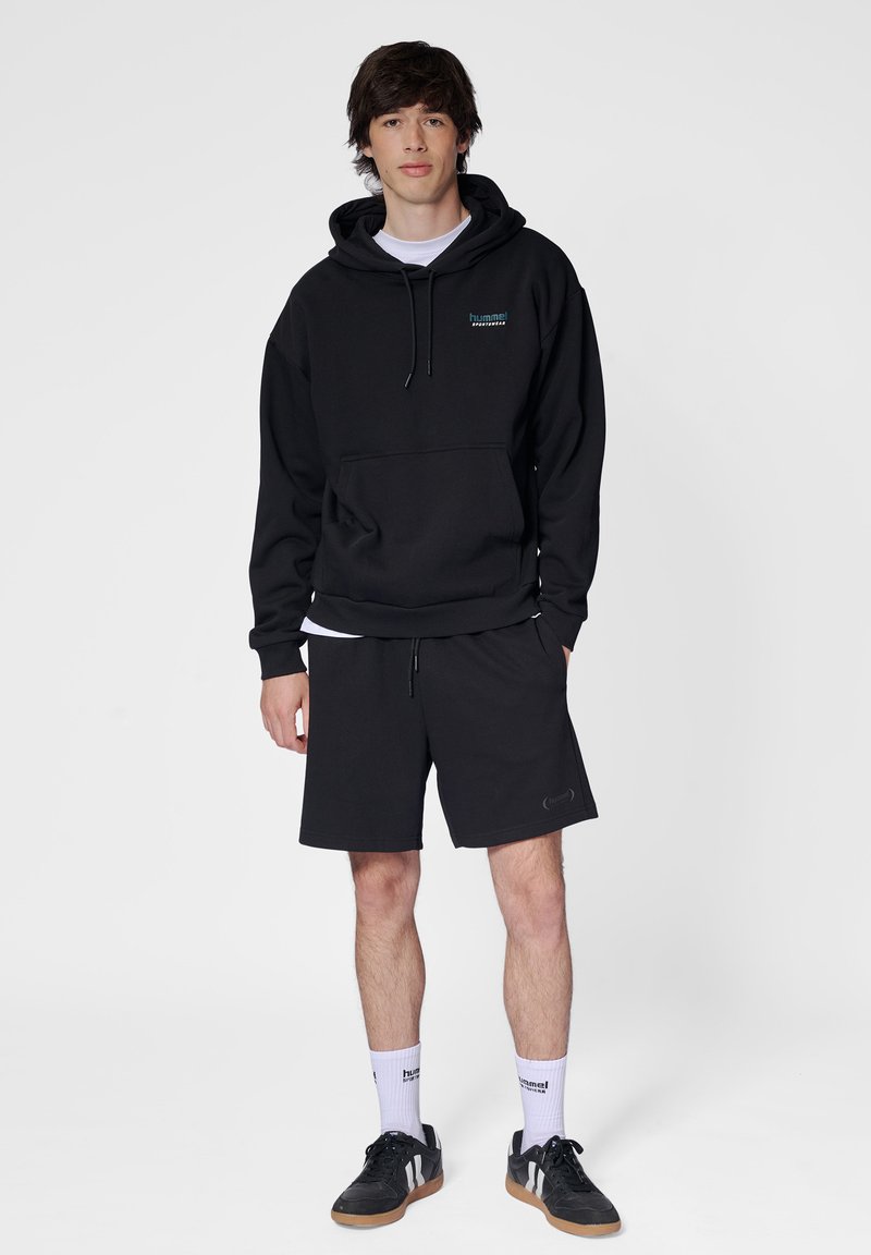 Zwarte pullover hoodie met een voorzak, gecombineerd met zwarte shorts. Model draagt een witte t-shirt eronder, zwarte sneakers en witte sokken.