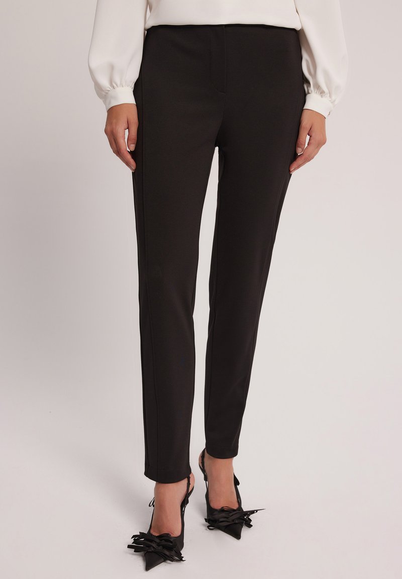 Czarne dopasowane spodnie o klasycznym kroju, z przednimi kieszeniami i gładką teksturą. W połączeniu z czarnymi czółenkami na szpilce z kokardą.