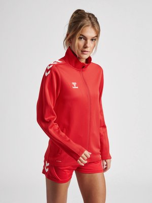 Femme portant une veste de sport rouge zippée et un short assorti avec des motifs chevrons blancs sur les manches et le short.