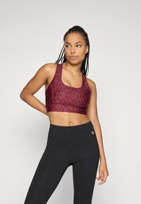 Kvinne i en rød og svart mønstret sports-bh og svarte tights, som står med den ene hånden på hoften og ser til siden.