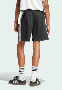 Shorts de sport noirs avec une taille élastique, des rayures blanches sur les côtés et une texture lisse. Associés à des baskets noires et des chaussettes blanches rayées.