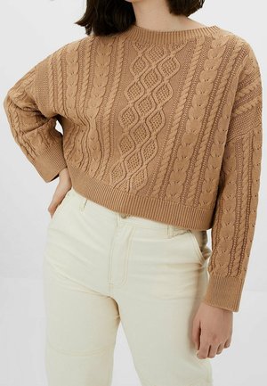 Kvinde iført en cropped kamelfarvet kabelstrikket sweater med lange ærmer og højttaljede off-white bukser, poserer med den ene hånd på hoften.