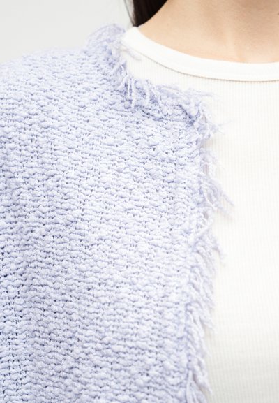 Κοντινό πλάνο ενός ατόμου που φοράει ένα υφαντό ένδυμα ανοιχτό μωβ με υφή και τελειώματα με κρόσσια πάνω από ένα λευκό ριμπ πλεκτό πουκάμισο.