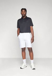 Čierne polo tričko s krátkym rukávom s logom, v kombinácii s bielymi šortkami. Model má na sebe biele tenisky a biele členkové ponožky. Postoj v stoji.