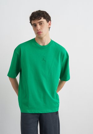 Młody mężczyzna z kręconymi włosami noszący zieloną oversizową koszulkę z subtelnym logo oraz ciemne spodnie, stojący na jednolitym białym tle.