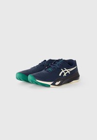 Chaussures de sport bleu marine avec une tige texturée, un talon vert et un détail de logo blanc. Elles présentent des lacets et une semelle en caoutchouc avec un motif antidérapant.