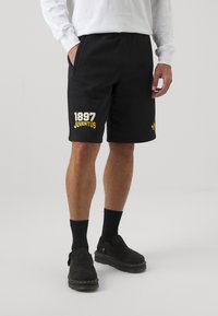 Black shorts with pockets, featuring a white and yellow graphic reading "1897 JUVENTUS" on the left leg. Soft, textured fabric.