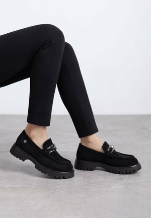 Mocasines de ante negro con suela de goma negra gruesa. Presentan detalles de hardware plateado en la parte superior, creando un acabado texturizado.