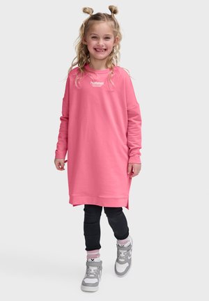 Smilende ung pige med blondt hår i to fletninger, iført en lang pink sweatshirt, sorte leggings, pinke sokker og grå sneakers, går fremad.