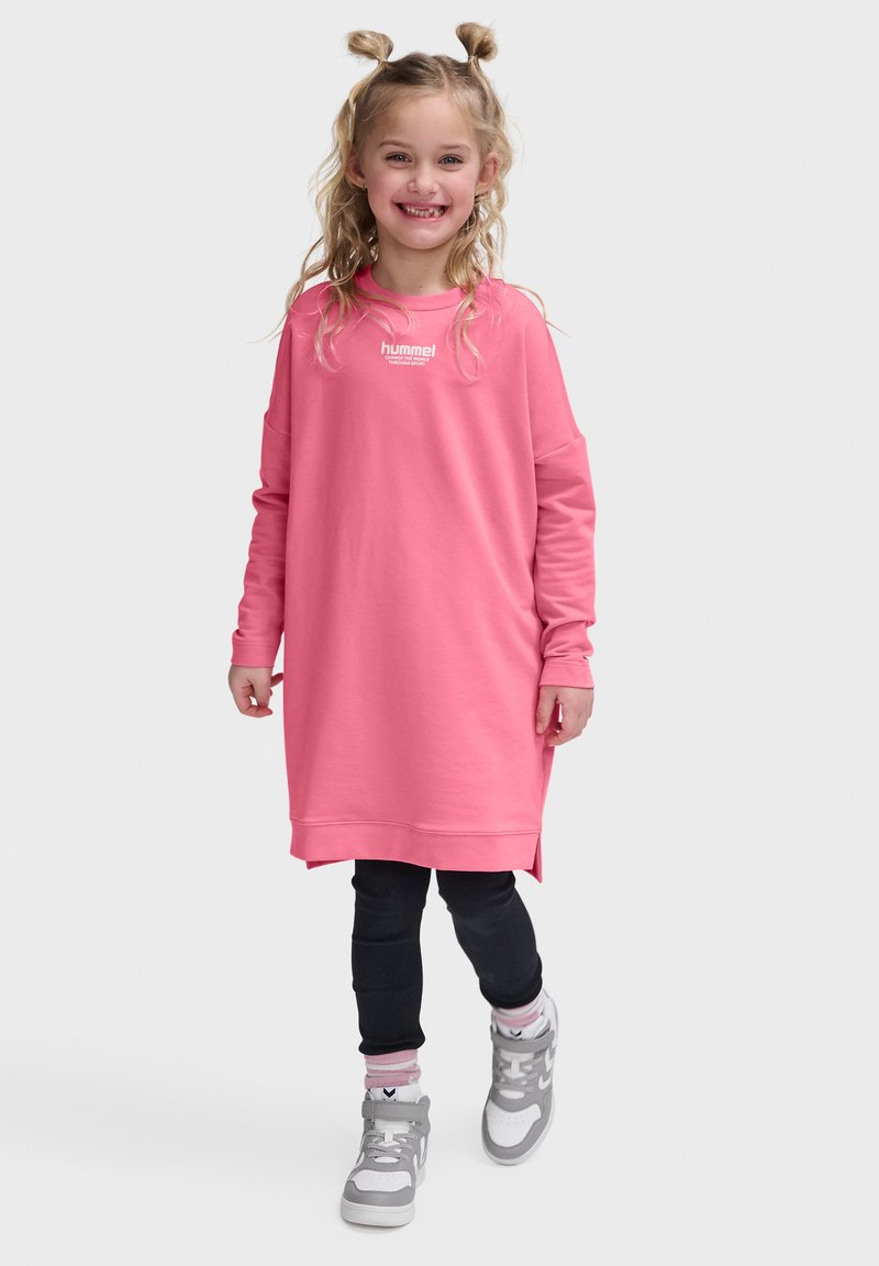 Smilende ung pige med blondt hår i to fletninger, iført en lang pink sweatshirt, sorte leggings, pinke sokker og grå sneakers, går fremad.