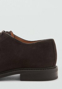 Zapato de ante marrón oscuro con textura suave, punta redondeada y un tacón de madera bajo y discreto. Cuenta con cordones y un forro interior en contraste de color caqui.