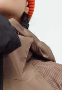 Person wearing an orange knit hat, tan coat with visible stitching, and a brown textured garment underneath, against a plain light background.