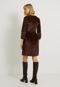 Vestido de estampado de leopardo marrón con mangas tres cuartos, cintura fruncida y sutiles bandas horizontales. Combinado con botas de cuero marrón hasta la rodilla.