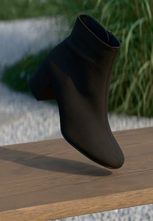 Botim negro até ao tornozelo feito de material suave, com um salto bloco e fecho de correr, exibido sobre uma superfície de madeira com relva verde ao fundo.