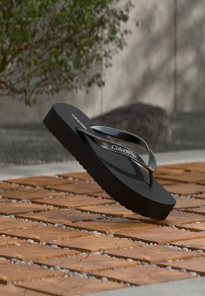 Fekete Calvin Klein platform papucs fényes pántokkal, fa csempén és kavicsos kültéri padlón, zöld bokor mellett pihenve.