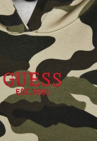 Haut à capuche avec un motif camouflage en vert et beige, doté d'un détail brodé rouge "GUESS EST. 1981" sur le devant. Tissu doux avec une coupe décontractée.