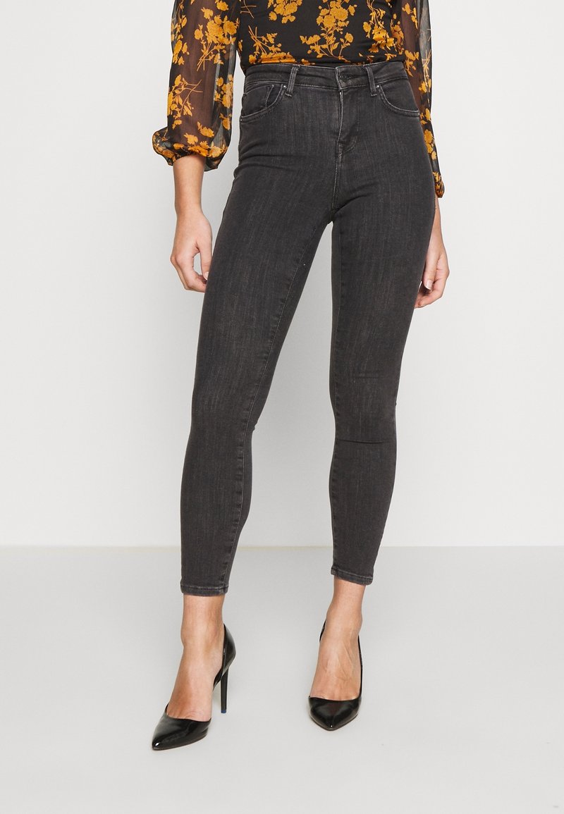 Jeans skinny de tiro alto en negro con una sutil textura de denim, diseño de cinco bolsillos y corte ajustado. Combinados con tacones altos negros.