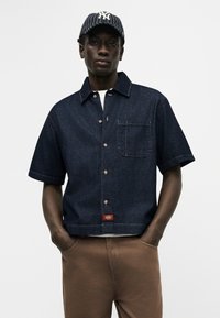 Άνδρας που φοράει σκούρο μπλε κοντομάνικο πουκάμισο από τζιν με τσεπάκι στο στήθος, καφέ παντελόνι και ασπρόμαυρο καπέλο μπέιζμπολ με ρίγες. Τα χέρια στις τσέπες.