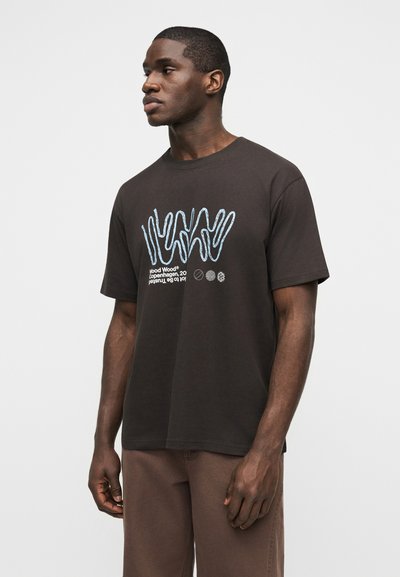 Μαύρος άνδρας που φοράει μαύρο γραφικό μπλουζάκι με μπλε κυματιστές γραμμές και λευκό κείμενο, συνδυασμένο με καφέ παντελόνι, στέκεται μπροστά σε απλό φόντο.