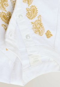 Chemise pour bébé blanche avec broderies en paisley doré et boutons transparents. L'étiquette indique "NEXT Baby". Tissu texturé avec de subtiles lignes verticales.