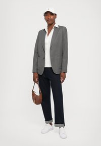 Gray tailored blazer, white button-up shirt, dark denim jeans, white sneakers, and a brown handbag. White cap with logo accents the look.