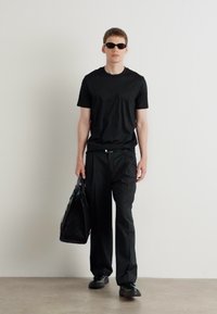 T-shirt nera a maniche corte e pantaloni neri larghi abbinati a scarpe nere. Il modello tiene in mano una borsa nera con trama e indossa occhiali da sole scuri.