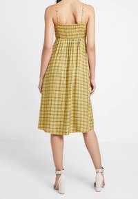 Vestido amarillo de cuadros vichy con cuerpo fruncido, tirantes finos, falda acampanada y diseño hasta la rodilla. Llevado con sandalias de tacón con tiras al tobillo de color claro.