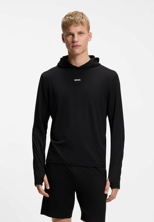Blonder Mann trägt einen schwarzen Hoodie mit langen Ärmeln und "BOSS"-Logo sowie schwarze Shorts, steht vor einem schlichten weißen Hintergrund.