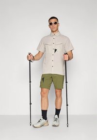 Ανοιχτό γκρι πουκάμισο με κοντό μανίκι, δύο τσέπες στο στήθος και μαύρο τρίγωνο διακοσμητικό, συνδυασμένο με σορτς ελιάς και μπαστούνια πεζοπορίας.