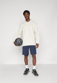 Polo de mangas compridas cinza claro com colarinho e três riscas nas mangas, combinado com calções azul-marinho. Bola de basquete preta na mão, usando ténis pretos e verdes.