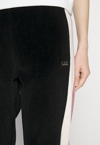 Detailní záběr na černé kalhoty s bílými a růžovými bočními pruhy a malým logem EA7 Emporio Armani blízko pravého boku, které nosí osoba.