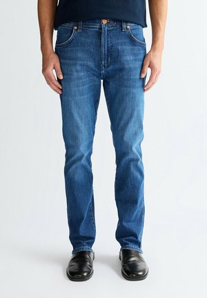 Persona che indossa jeans blu slim fit e scarpe di pelle nere, in piedi davanti a uno sfondo chiaro e semplice.