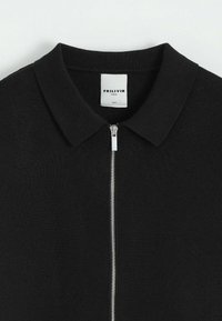 Polo noir zippé avec un tricot texturé, doté d'un col doux et d'une fermeture éclair en argent, portant l'étiquette "FRILVIN" à l'intérieur. Étiquette de taille visible.