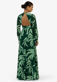 Robe maxi verte avec imprimé feuilles, manches longues, dos ouvert et un lien à la taille. Fabriquée en tissu léger avec une texture lisse.