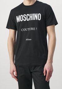 Man som bär en svart Moschino-t-shirt med vit text "MOSCHINO COUTURE! Milano" och svarta byxor, står med armarna avslappnat vid sidorna.