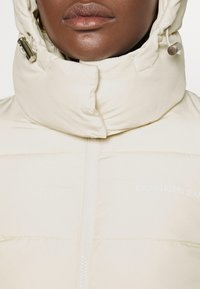 Giubbotto imbottito beige chiaro con colletto alto, dotato di zip frontale, dettagli con logo e una superficie liscia e trapuntata.