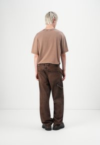 Persona con cabello rubio corto que lleva una camiseta marrón holgada, pantalones cargo marrón oscuro y zapatos negros gruesos, de pie contra un fondo blanco liso.