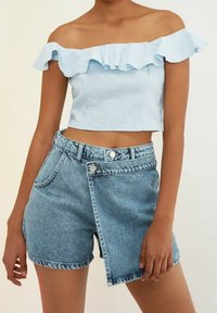 Top corto azul claro sin hombros con ribete de volantes; combinado con shorts de mezclilla de talle alto en un lavado claro desgastado con cierre de botones.