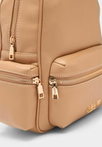 Mochila de piel sintética color tan con cremalleras de tono dorado, dos bolsillos frontales con cremallera y detalles sutiles de costura. Acabado suave y texturizado.