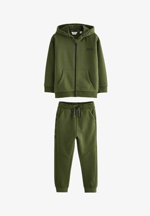 Zöld kapucnis pulóver és melegítőnadrág szett, puha anyagból készült, húzózsinóros kapucnival és bordázott mandzsettákkal, fehér sportcipővel párosítva.
