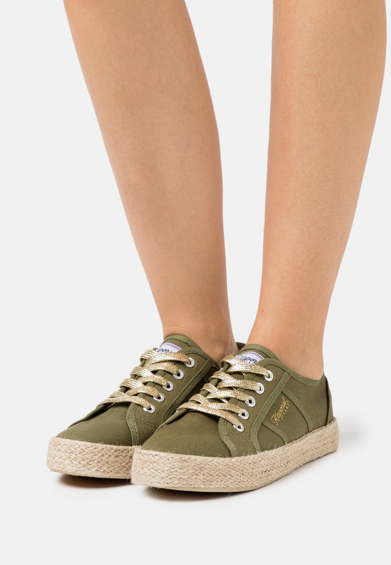 Pieds portant des baskets basses en toile vert olive avec des semelles beiges tissées épaisses et des lacets parsemés d’or sur fond blanc.