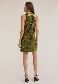 Rochie verde strălucitoare din paete, fără mâneci, cu un design ajustat, având un decolteu rotund și o lungime mini, asociată cu tocuri înalte negre.