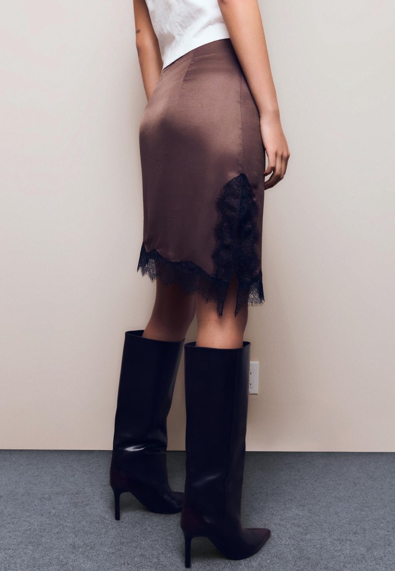 Femme portant une jupe en satin marron avec un bord en dentelle noire et des bottes à talons noires montant jusqu'aux genoux, debout contre un mur beige.