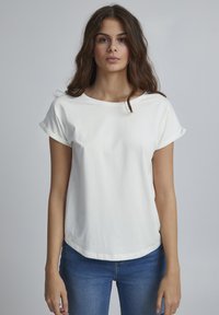 Balta medvilnė t-shirt su užverčiamais rankovėmis, apvaliu kaklu ir laisvu siluetu. Dėvimi kartu su mėlynomis džinsinėmis kelnėmis, kurios pasižymi lygiu paviršiumi ir minimaliai išsiskiriančiu dizainu.