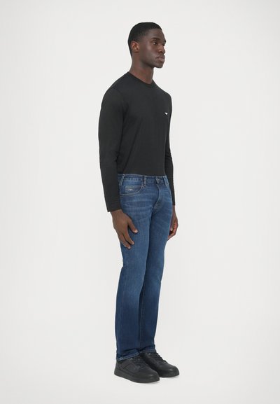 Camisa negra de manga larga, jeans de mezclilla azul y zapatillas negras. Los jeans tienen un diseño desgastado con un detalle distintivo en el bolsillo.