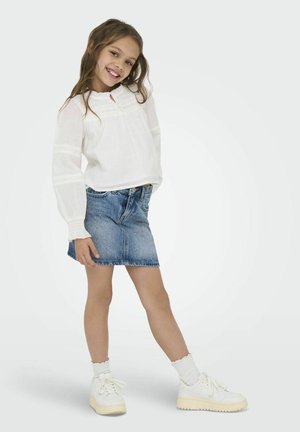 Bela bluza z dolgimi rokavi s čipkastim robom, v kombinaciji z modrim denim krilom. Ima bele superge in rebraste bele nogavice.