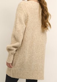 Lång, beige stickad kofta med avslappnad passform, ribbade manschetter och fåll; mjuk textur med ett subtilt melerat mönster, når till mitten av låret.