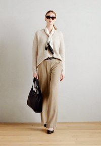 Model in a beige sweater, black scarf, and striped beige trousers, holding a black tote bag. Features large black sunglasses and pointed shoes.