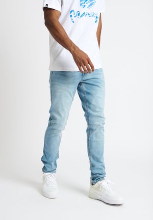 Light blue denim jeans with a slim fit. Paired with a white t-shirt featuring blue graphic text. White sneakers complete the outfit.